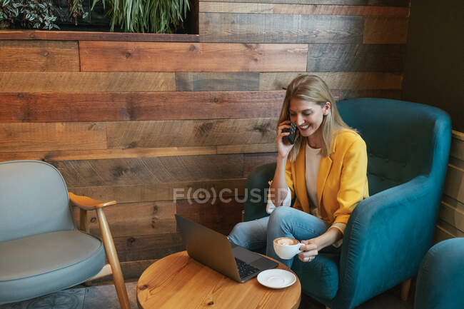 Alto ángulo de risa freelancer femenina adulta en ropa casual que se centra en la pantalla del ordenador portátil y escribir mientras está sentado en un sillón azul suave en la mesa redonda de madera con taza de bebida caliente y hacer una llamada telefónica en la cafetería moderna - foto de stock