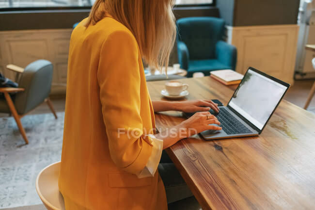 Cropped unrecognizable young female freelancer in casual clothes having tasty hot drink while sitting at wooden table and using laptop in light modern cafe — Stock Photo