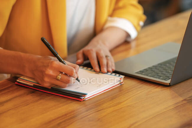Низкий угол растениеводства деловая женщина в элегантной красочной одежде, пишущая ручкой в ноутбуке, сидя за деревянным столом и используя ноутбук в светлом современном офисе — стоковое фото