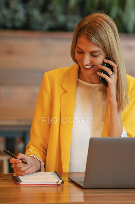 Adulta mujer de negocios llena de alegría en ropa formal mirando hacia otro lado y riendo mientras habla por teléfono sentado en la mesa de madera con el ordenador portátil y tomando notas en el cuaderno en la oficina contemporánea - foto de stock