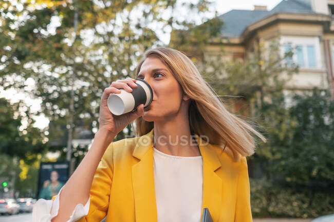 From below trendy slim blond businesswoman looking away in elegant vivid yellow jacket and jeans with digital tablet walking alone against exteriors of residential multistory buildings and cars parked on street in downtown — Stock Photo
