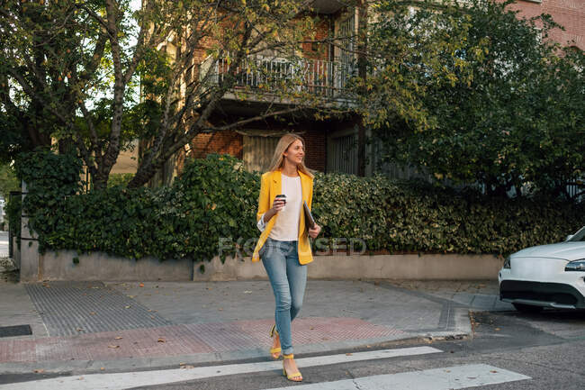 Модна струнка блондинка-бізнесмен, дивлячись в елегантний яскраво-жовтий піджак і джинси з цифровим планшетом, що йде один навпроти екстер'єрів житлових багатоповерхових будівель і автомобілів, припаркованих на вулиці в центрі міста — стокове фото