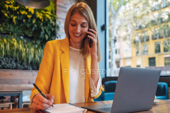 Adulto mulher de negócios muito feliz em desgaste formal olhando para longe e rindo ao falar ao telefone sentado à mesa de madeira com laptop e tomar notas no notebook no escritório contemporâneo — Fotografia de Stock