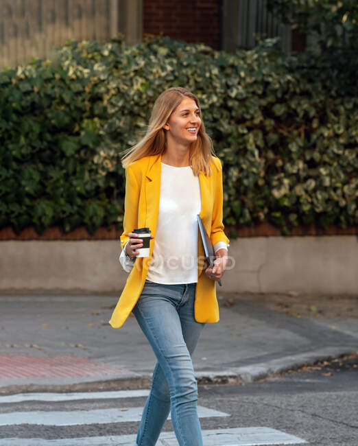 Модна струнка блондинка-бізнесмен, дивлячись в елегантний яскраво-жовтий піджак і джинси з цифровим планшетом, що йде один навпроти екстер'єрів житлових багатоповерхових будівель і автомобілів, припаркованих на вулиці в центрі міста — стокове фото