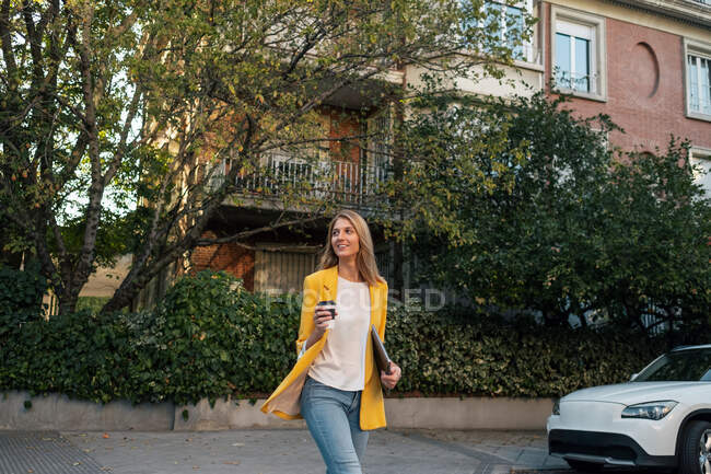 Модна струнка блондинка, дивлячись в елегантний яскраво-жовтий піджак і джинси з цифровим планшетом, що йде один проти екстер'єрів житлових багатоповерхових будівель і автомобілів, припаркованих на вулиці в центрі міста — стокове фото