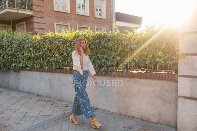Вид збоку позитивної розслабленої блондинки-жінки в повсякденному одязі, що йде по вулиці і посміхається, говорячи на смартфоні біля паркану, покритого зеленим скелелазінням рослини — стокове фото