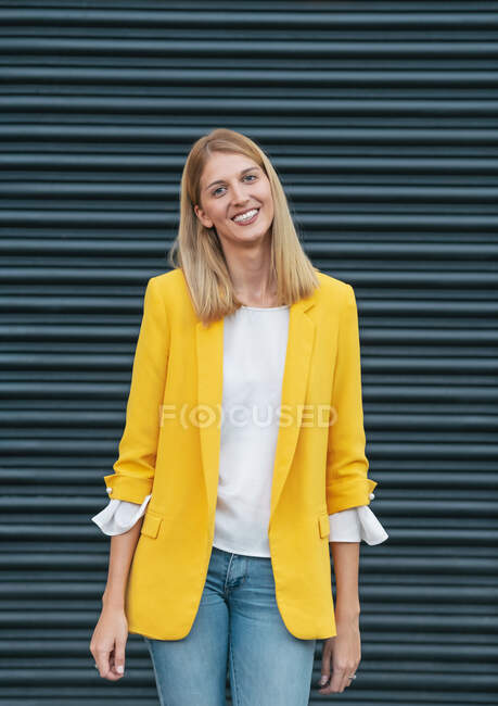 Happy young blond female in bright yellow jacket and jeans smiling looking at camera while standing on street against blurred striped wall in city — Stock Photo