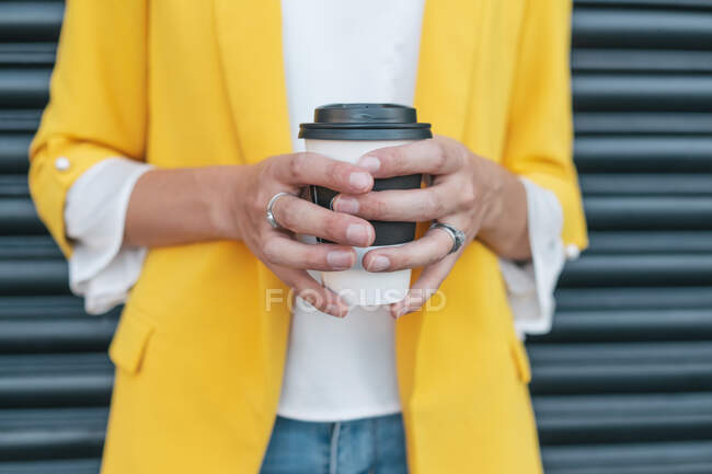 Low angle of crop trendy relaxed female in vibrant yellow jacket holding takeaway cup of yummy aromatic coffee against striped wall on street in city — Stock Photo