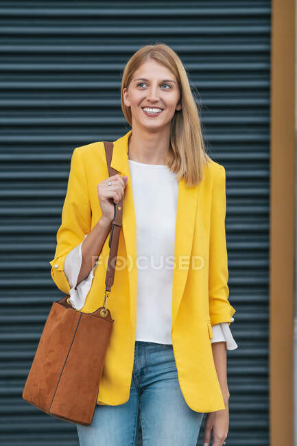Joyeux jeune femme blonde dans une veste jaune vif et un jean avec sac à main marron sur l'épaule souriant tout en se tenant dans la rue contre un mur rayé flou en ville — Photo de stock