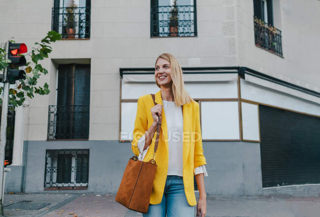 Joyeux jeune femme blonde en veste jaune vif et un jean avec sac à main marron sur l'épaule souriant loin en attendant de traverser sur le feu de circulation dans la rue dans la ville — Photo de stock