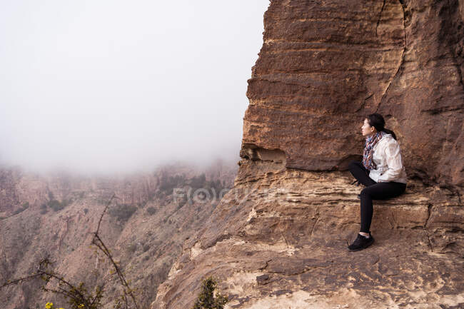 Vista lateral da fêmea asiática em roupas casuais sentadas em um penhasco áspero e olhando para longe no dia nebuloso nas montanhas — Fotografia de Stock