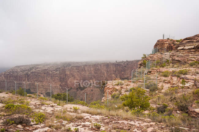 Breathtaking view of rough rocky cliff with green shrubs and metal barrier located in mountainous terrain on misty day — Stock Photo