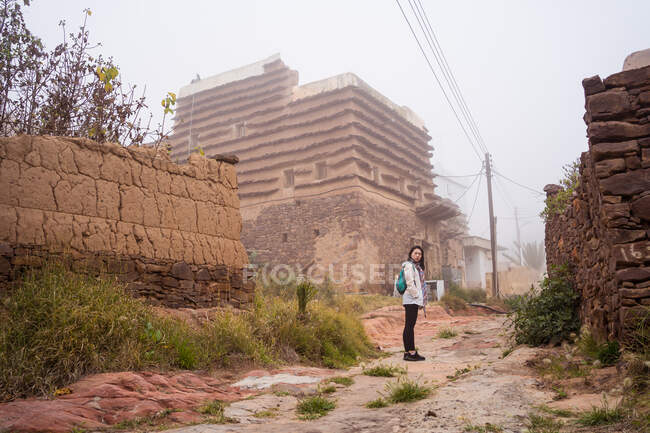 Side view of ethnic female traveler looking at camera over shoulder while standing near weathered buildings of old district on gray day in town — Stock Photo