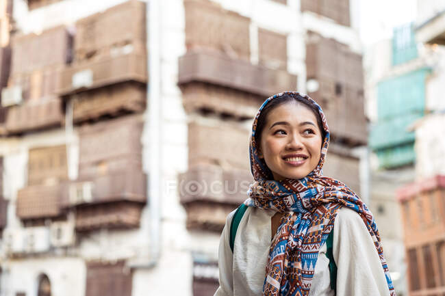 Jeune femme ethnique en foulard souriant et détournant les yeux tout en se tenant debout sur un fond urbain flou dans la ville de Jeddah en Arabie Saoudite — Photo de stock
