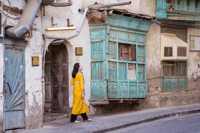 Vue latérale d'une femme ethnique marchant le long d'un immeuble résidentiel en pierre minable dans un quartier pauvre de la ville de Jeddah en Arabie Saoudite — Photo de stock