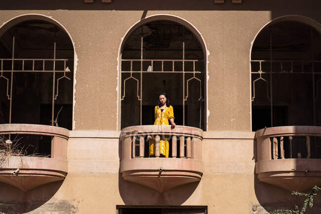 Niedriger Winkel einer jungen ethnischen Frau in gelbem Kleid, die auf einem kleinen gewölbten Balkon eines alten Steingebäudes in Dschidda in Saudi-Arabien steht — Stockfoto