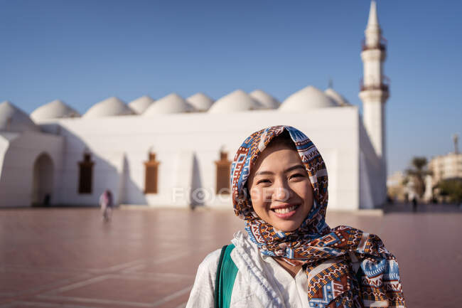 Happy young Asian female in traditional headscarf smiling and looking at camera while standing on square against Islam mosque in Jeddah city in Saudi Arabia — Stock Photo