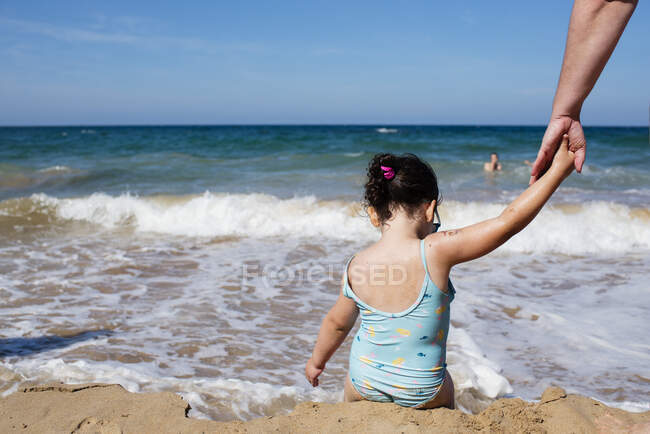 Rückansicht des kleinen anonymen Mädchens im Badeanzug, das die Hand der Eltern hält, während es am nassen Sandstrand sitzt und warmes Wasser vor dem Hintergrund der majestätischen Meereslandschaft genießt — Stockfoto