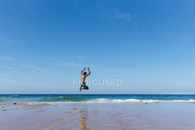 Вид ззаду на невпізнаваного чоловіка в купальнику, який насолоджується літнім відпочинком і стрибає у морську воду на блакитне небо в сонячний день — стокове фото