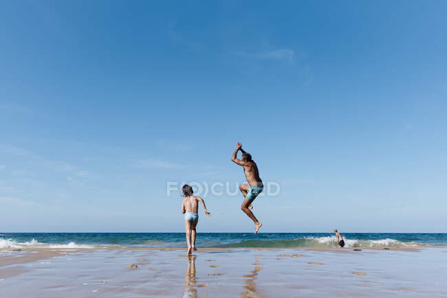Обратный вид неузнаваемого человека с маленьким сыном, бегущим и прыгающим в морскую воду, веселясь вместе во время летних каникул на песчаном пляже — стоковое фото