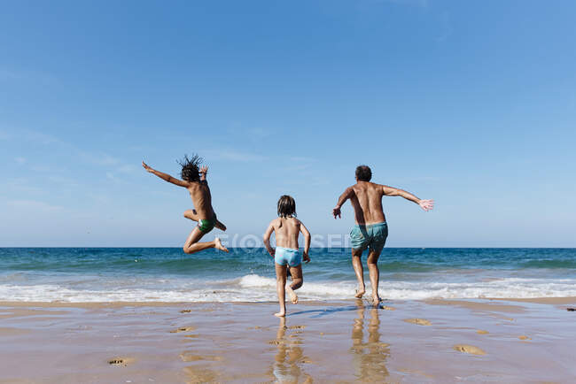 Вид ззаду на невпізнаваного чоловіка з маленькими синами біжить і стрибає у морську воду, розважаючись разом під час літніх канікул на піщаному пляжі — стокове фото
