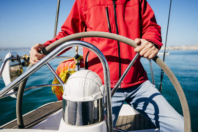 Crop unrecognizable man in red jacket and jeans standing at steering wheel while sailing in sea on modern vessel — Stock Photo