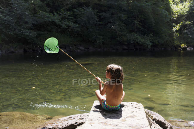 Вид ззаду без сорочки хлопчик сидить на камені і ловить рибу з сіткою, проводячи літні канікули на березі річки — стокове фото