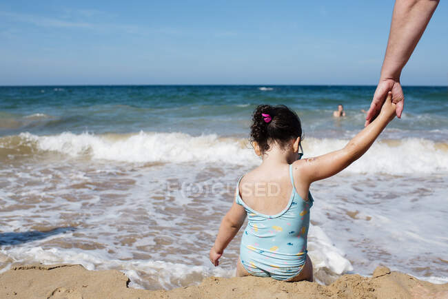 Back view of little anonymous girl in swimsuit holding hand of crop parent while sitting on wet sandy beach and enjoying warm water on background of majestic seascape — Stock Photo