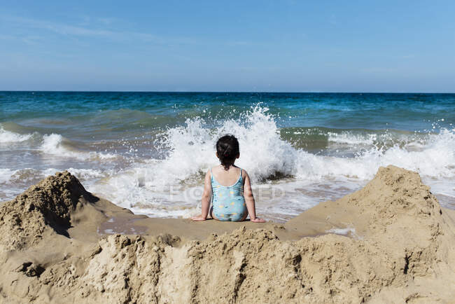 Обратный вид на неузнаваемую маленькую девочку в купальнике, сидящую на песчаном пляже против морских волн и наслаждающуюся летним отдыхом в солнечный день — стоковое фото