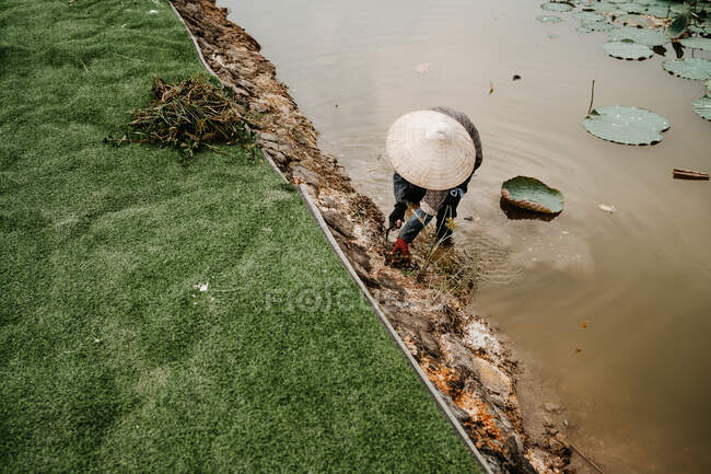 Высокий угол неузнаваемого занятого фермера в соломенной шляпе, стоящего в воде грязной реки и культивирующего почву мотыгой — стоковое фото