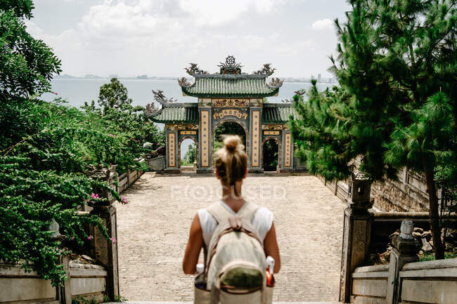 De cima vista lateral do viajante encantado fêmea com mochila passeando no jardim tropical perto do templo religioso e desfrutando de paisagem majestosa — Fotografia de Stock