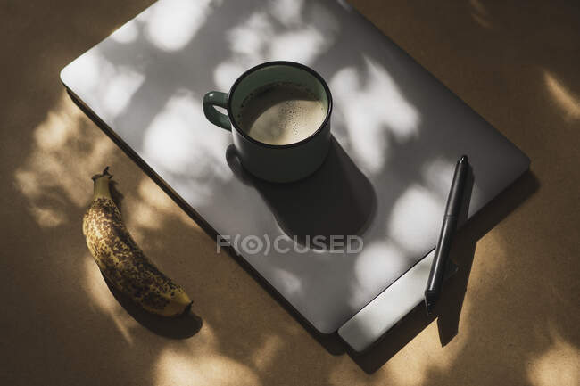 Caneca de café em comprimido gráfico com caneta e banana madura à luz do sol — Fotografia de Stock