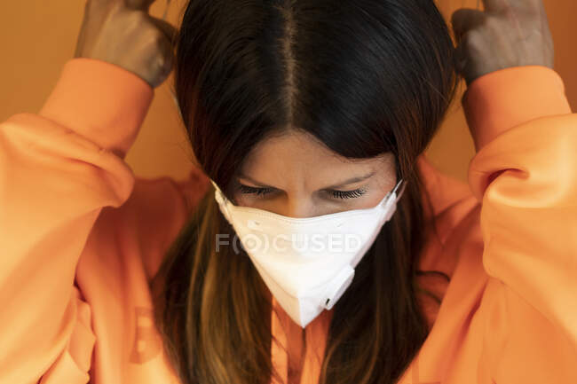 Jovem fêmea em jaqueta laranja casual colocando máscara protetora branca contra fundo laranja — Fotografia de Stock