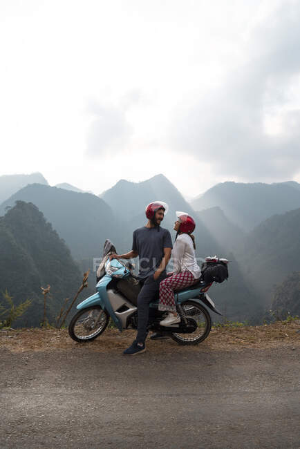 Vista laterale della coppia felice di viaggiatori seduti sulla moto parcheggiata sul ciglio della strada mentre si guardano sullo sfondo della catena montuosa — Foto stock