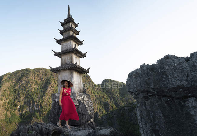 Femme voyageuse portant une robe d'été et un chapeau conique profitant des vacances d'été sur le dessus de la grotte Mua tout en se tenant près de la tour bouddhiste traditionnelle — Photo de stock