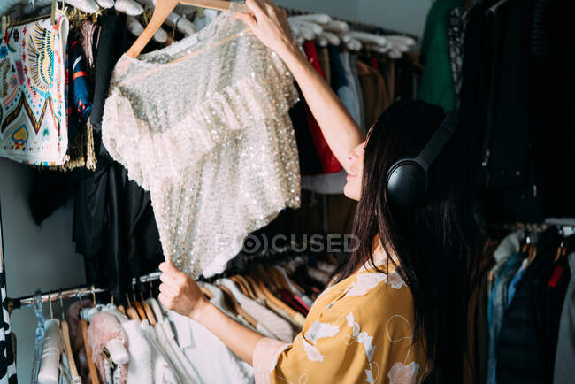 Вид збоку чарівна брюнетка в шовковій сукні стоїть в гардеробі в квартирі і вибирає вбрання, слухаючи музику з навушниками — стокове фото