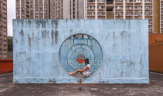 Feminino em vestido de verão sentado na base de concreto de instalação da cidade incomum em forma de passagem com buracos redondos e olhando para longe — Fotografia de Stock
