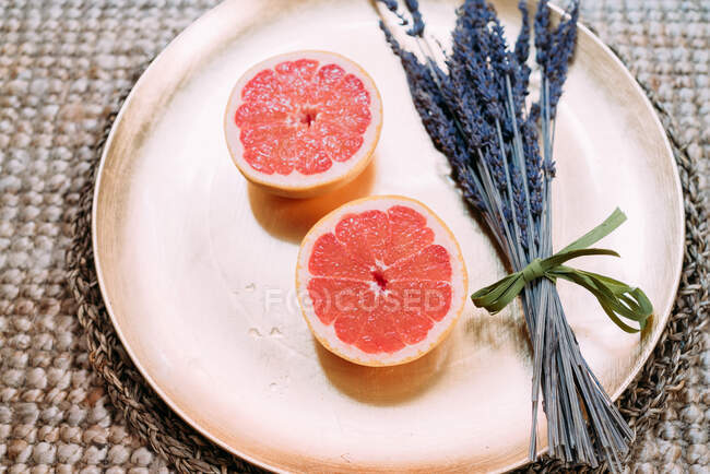 Сверху золотая тарелка с половиной свежесочного грейпфрута рядом с небольшим букетом сушеной лаванды, расположенной на плетеном ротанговом подставке на ковре — стоковое фото