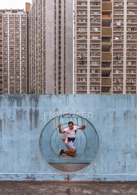 Homem encantado em shorts de verão e t-shirt pulando perto de parede de concreto com buracos de túnel localizado no pátio cercado por edifícios de condomínio — Fotografia de Stock