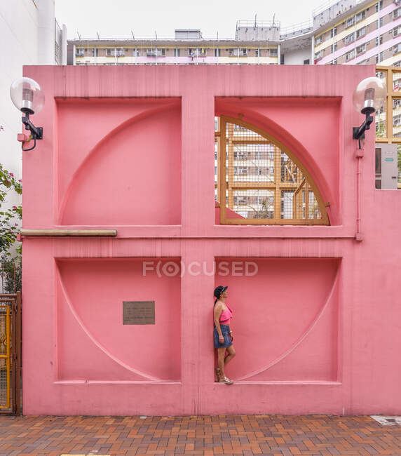 Vista lateral de la mujer en traje de verano de pie cerca de la pieza de pared de color rosa en forma de ventana en el barrio de Shek Kip Mei - foto de stock
