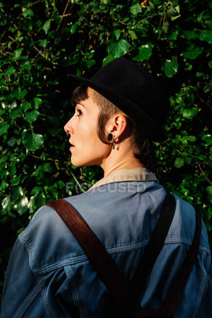 Back view of young stylish lady with stylish black hat standing near blooming bushes and looking away — Stock Photo