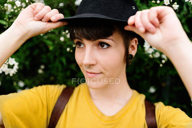 Señorita joven y positiva con estilo en camisa amarilla ocasional y sombrero negro elegante que se encuentra cerca de arbustos florecientes y mirando hacia atrás. - foto de stock