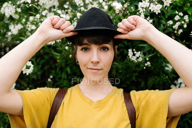 Позитивна молода стильна леді в повсякденній жовтій сорочці і стильний чорний капелюх, що стоїть біля квітучих кущів і дивиться на камеру — стокове фото