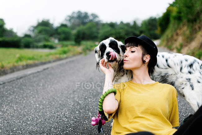 Positive junge Frau in Freizeitkleidung und schwarzem Hut, die sich beim Spaziergang im Park mit Hund auf den Boden legt — Stockfoto