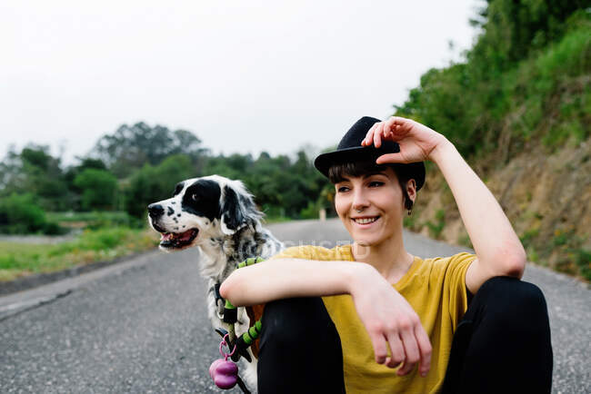 Jeune femme positive en vêtements décontractés et chapeau noir reposant sur le sol avec chien pendant la promenade dans le parc — Photo de stock