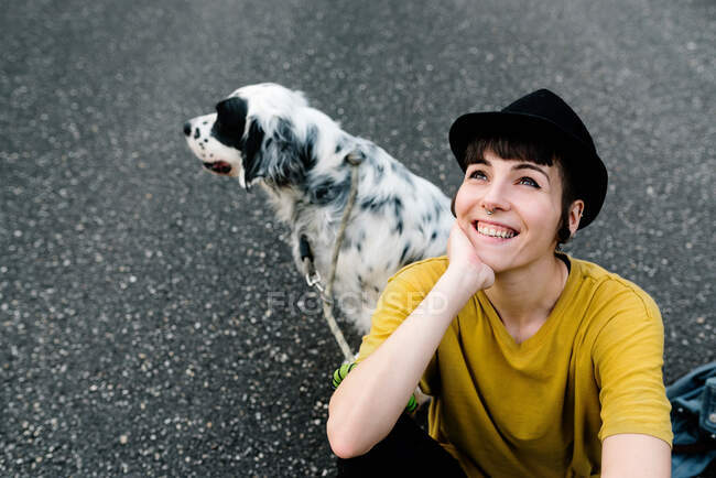 De cima positivo jovem senhora em roupas casuais e chapéu preto descansando no chão com cão durante a caminhada no parque — Fotografia de Stock
