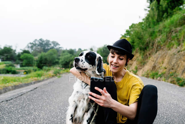 Веселая юная леди в повседневной одежде и шляпе сидит на земле с мобильным телефоном и делает селфи с собакой во время прогулки по улице — стоковое фото