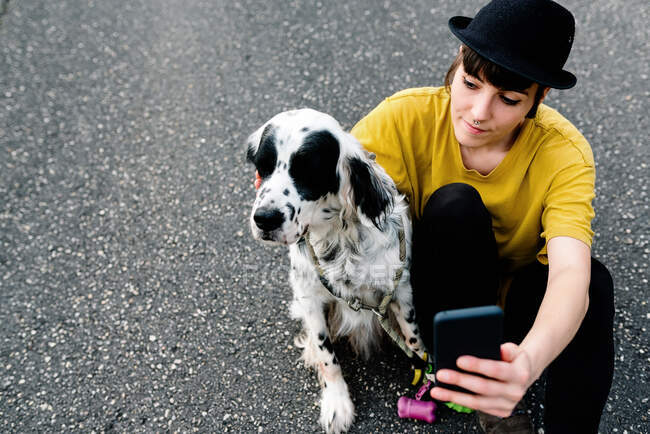 Jovem senhora em roupas casuais e chapéu sentado no chão com telefone celular e tomar selfie com cão durante a caminhada na rua — Fotografia de Stock