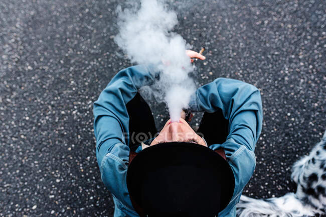 From above view of unrecognizable female in stylish black hat and casual clothes sitting on asphalt road and smoking cigarette while resting during walk — Stock Photo