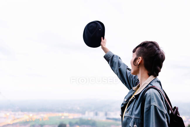 Side view of unrecognizable young lady in casual clothes with backpack and hat standing on hill with raised hand and admiring cityscape while spending time in evening — Stock Photo